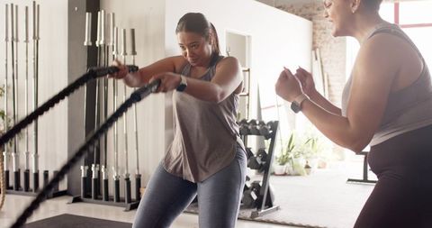 Trainer coaching client through battle ropes workout in sunlit fitness studio