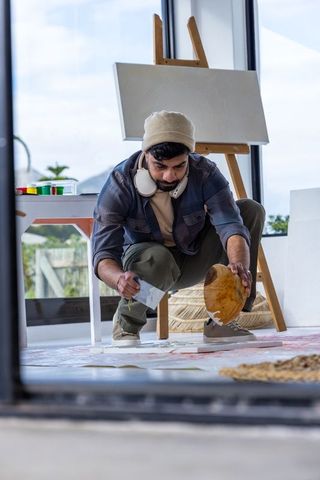 Asian man pouring paint from wooden bowl while crouching in sunlit art studio