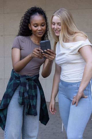 Diverse female friends sharing smartphone, smiling and leaning against tiled wall, casual urban