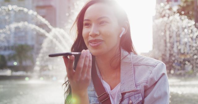 Young woman using smartphone to record voice message while sitting in urban park with city fountain. Ideal for themes like communication technology, outdoor lifestyles, relaxation, modern connectivity, and urban living.
