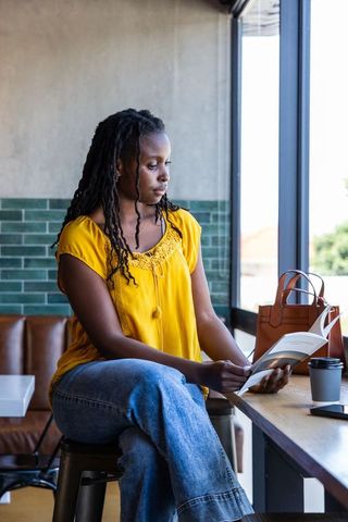 African american woman sitting at cafe counter reading book with coffee and leather tote