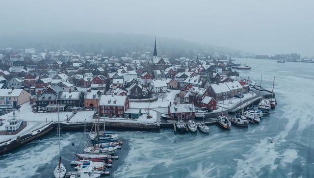Snow-covered coastal town nestled around frozen harbor with boats and church steeple