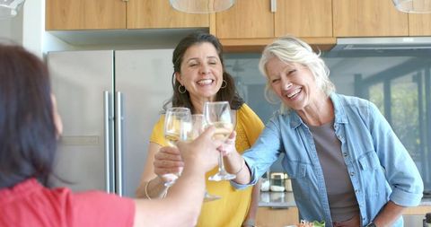 Mature friends clinking white wine glasses in bright kitchen, laughing, celebrating