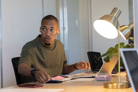 African american man reaching for smartphone while working on laptop in coworking space