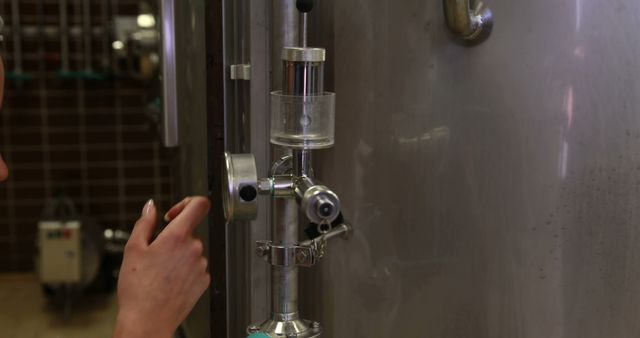 Close-up of a hand operating stainless steel brewery equipment, focusing on the control valves and machinery. Useful for illustrating brewing, beer production processes, industrial equipment, and manufacturing settings.