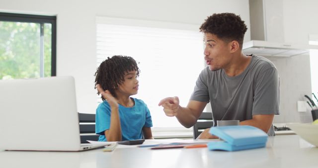 Father teaching son at home, creating bonding experience through learning. Both are engaged with open laptop and study materials on table. Great for concepts of modern parenting, online education, and family bonding. Ideal for promoting family values, homeschooling, and educational tools.