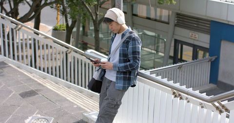 Asian commuter leaning on overpass railing checking phone with headphones, takeaway coffee