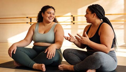 Smiling plus-size women chatting after workout in sunlit dance studio with yoga mats