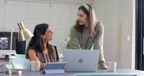 Two diverse women collaborating and brainstorming at shared workspace with laptop and tablet