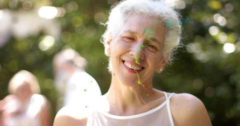 Smiling senior woman wearing white top with colorful paint specks in sunlit garden closeup