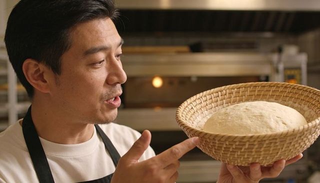 Asian baker holding banneton with risen dough explaining proofing process in artisan bakery