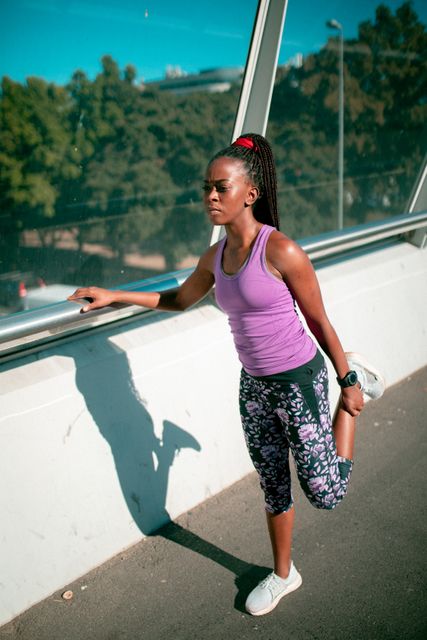 African American woman stretching her leg on a bridge at sunset. She is wearing sportswear and appears focused on her fitness routine. This image is ideal for promoting healthy living, fitness programs, outdoor exercise, and active lifestyle campaigns. It can also be used in articles or advertisements related to workout routines, sportswear, and urban fitness activities.