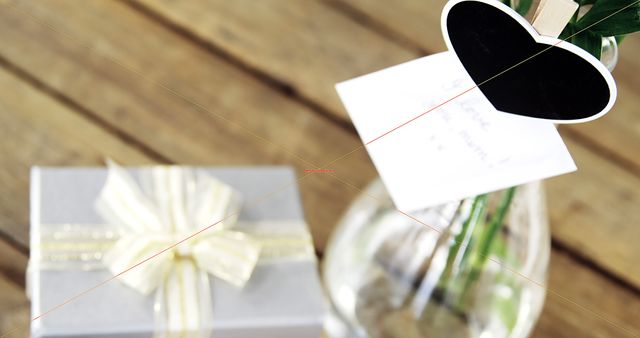 Close-up view of a beautifully wrapped gift with a bow on a wooden table. A heartfelt note is attached to a heart-shaped card placed inside a glass vase. Suitable for themes related to celebrations, love, gifting, and special occasions. Ideal for use in blogs, websites, presentations, and advertisements regarding holidays, birthdays, anniversaries, or romantic gestures.