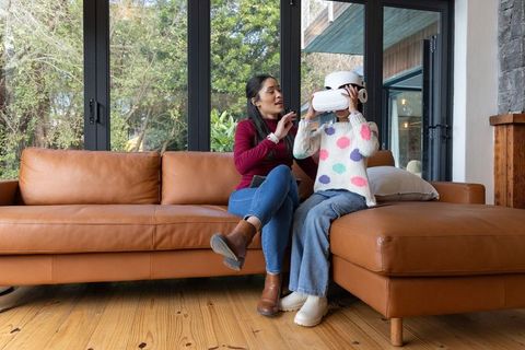 Asian mother helping child try virtual reality headset on cozy leather sofa at home
