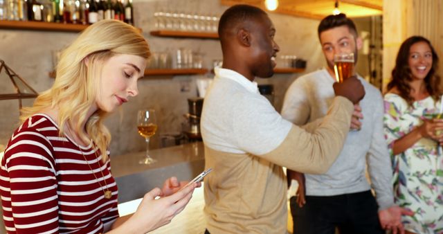 Diverse group of friends socializing at a bar. One person using a smartphone while others are talking and enjoying drinks. Useful for themes related to social interactions, nightlife, modern technology, friendship, and leisure activities.
