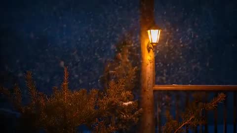Snow Falling on Wooden Deck Lantern Casting Warm Light at Night