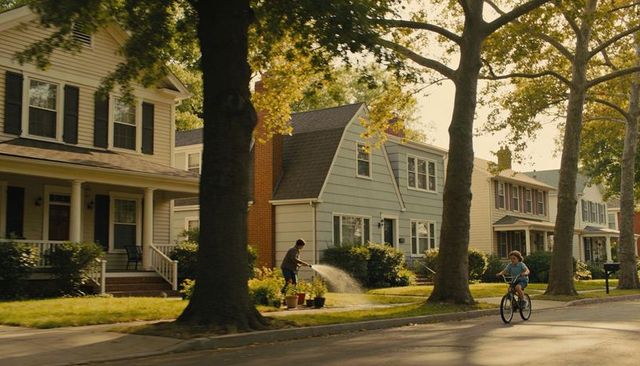 Boy riding bicycle past man watering lawn on tree-lined suburban street at sunset