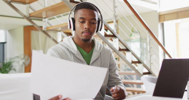 Young man wearing headphones and concentrating on work with a laptop and papers at a modern home office. Perfect for illustrating remote work, e-learning, study habits, or modern workplaces. Useful for promoting work-from-home setup ideas, productivity apps, and co-working spaces.