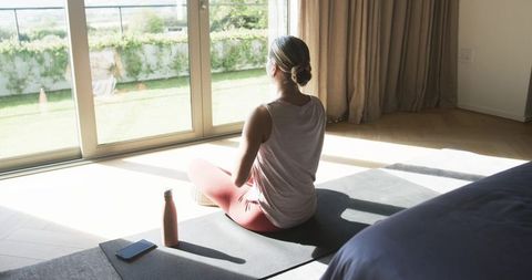Woman meditating on yoga mat by sunlit sliding doors at home for mindfulness and wellness