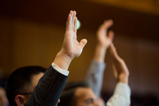 Business executives engaging actively in a conference by raising hands. Suitable for use in contexts involving corporate events, seminars, business meetings, teamwork, professional engagement, and audience participation.
