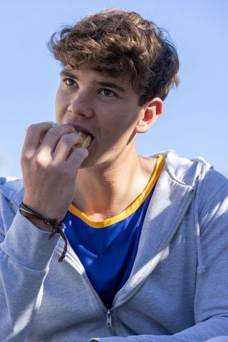 Teenager eating sandwich outdoors in athletic jersey and gray hoodie, casual snack