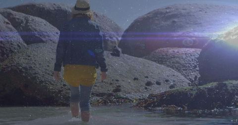 Woman in Boots Navigating Shallow Ocean Near Boulders