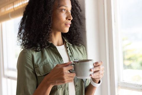 African american woman gazing out window holding ceramic mug in sunlit home morning mindfulness