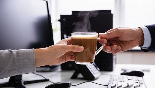 Colleagues passing steaming glass coffee mug over desk during quick office break moment