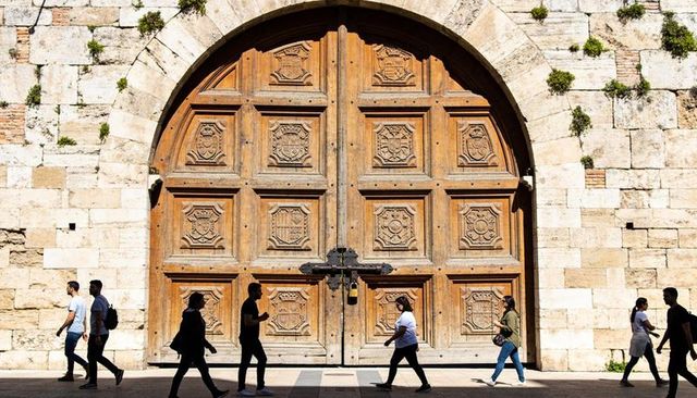 Sunlit carved wooden double door with iron bar padlock at stone archway, pedestrians walking