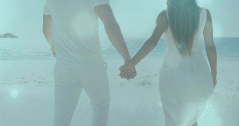 Walking hand in hand toward ocean in white clothing with soft sun flare on pastel beach