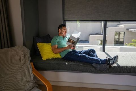 Mature asian man relaxing on sunny window seat reading paperback with yellow cushion