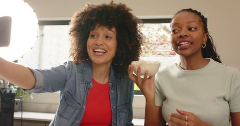 Two female creators filming cooking reel while smiling and holding bowl in bright kitchen