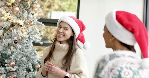 Friends wearing santa hats laughing by flocked christmas tree in cozy home interior