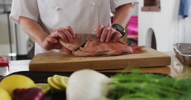 Chef in white uniform preparing fresh fish on a wooden cutting board in kitchen, focus on hands and ingredients; ideal for content on culinary techniques, cooking classes, fresh food preparation, healthy eating and professional kitchens.