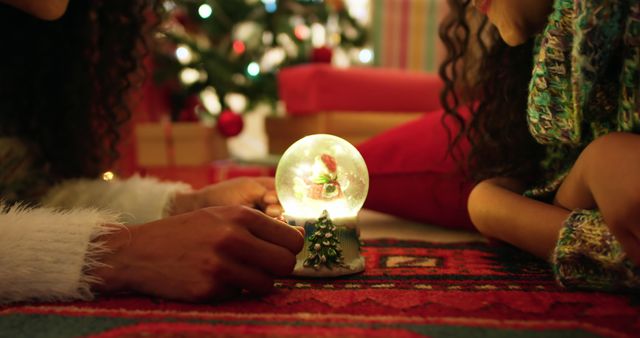 Close-up of family members admiring a snow globe with Christmas decorations in the background. Warm, cozy winter scene with Christmas tree and presents. Ideal for themes of family, holidays, Christmas cheer, and festive home interiors. Perfect for holiday cards, festive advertisements, and articles on holiday traditions and celebrations.