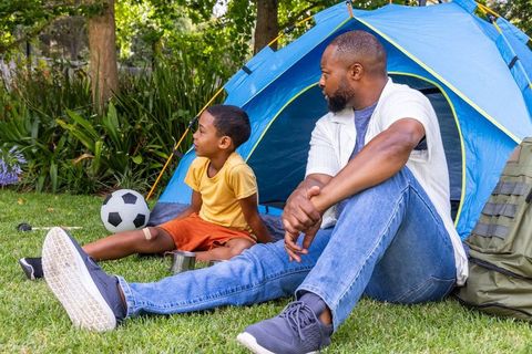 African american father and son relaxing by blue tent in backyard camping with soccer