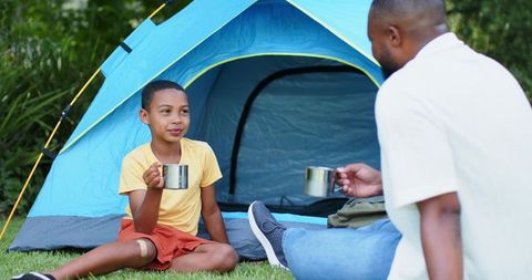 African american father and son camping sharing hot drinks family bonding by blue tent