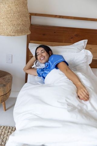 Smiling woman stretching in blue pajamas on minimalist white bed with natural wood
