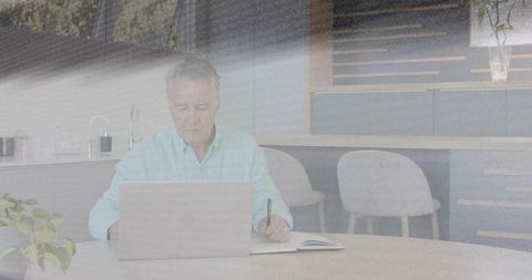 Mature man working on laptop while writing notes at round table in bright modern kitchen
