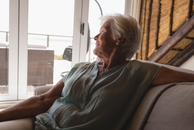 This image depicts a senior woman sitting on a couch in a living room, looking thoughtfully out the window. The natural light and serene expression suggest a moment of peaceful contemplation. This image can be used for topics related to aging, retirement, mental health, relaxation, and home life.