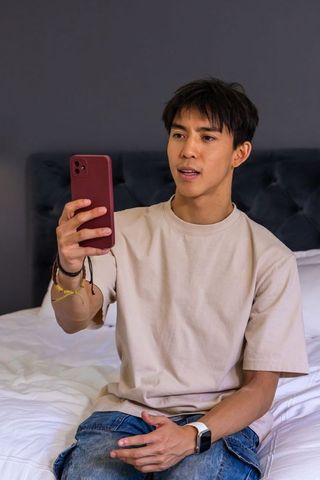 Asian man sitting on bed holding smartphone and speaking, casual bedroom lifestyle portrait