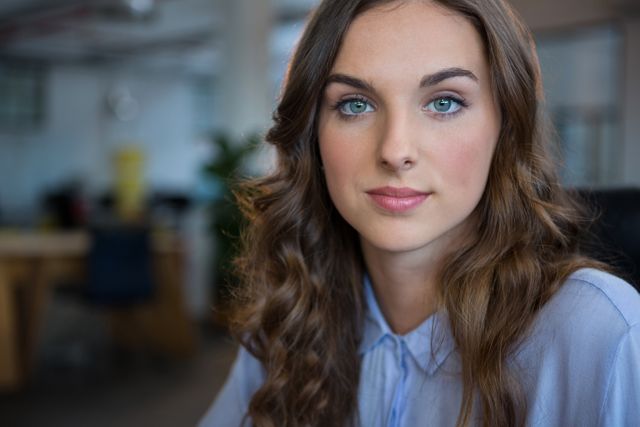 This stock image features a young businesswoman with wavy hair and blue eyes, smiling confidently in a modern office environment. Perfect for use in corporate presentations, websites, marketing materials, and articles focusing on professional development, leadership, and career success.