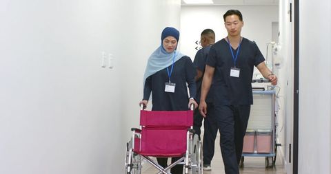 Healthcare team walking corridor, nurse in headscarf pushing wheelchair, colleague with cart