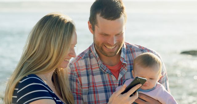 Happy caucasian mother and father holding baby and using smartphone in sun by the sea. Communication, parenthood, family, care, wellbeing, vacations and travel, unaltered.