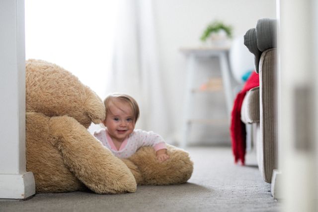 Happy caucasian baby daughter lying on floor with giant teddy bear in living room and smiling. at home in isolation during quarantine lockdown.