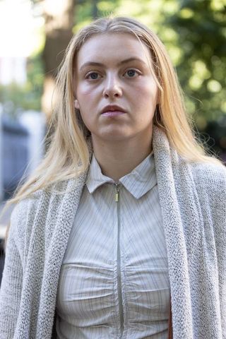 Young blonde woman standing on tree-lined sidewalk wearing cardigan and striped blouse