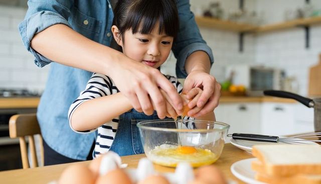 Parent guiding child cracking egg in home kitchen bonding over cooking and learning