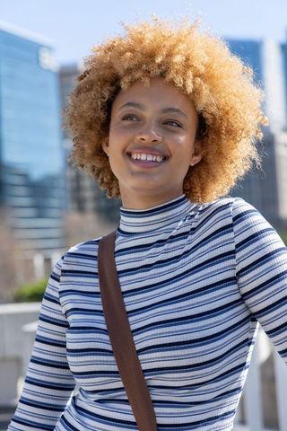 Smiling african american woman with curly blonde hair posing in urban daylight in striped top