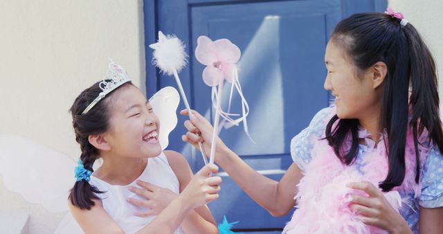Two Asian girls are enjoying a playful moment in costumes, with one wearing fairy wings and the other holding a wand, with copy space. Their laughter and imaginative play highlight the joy and creativity of childhood.