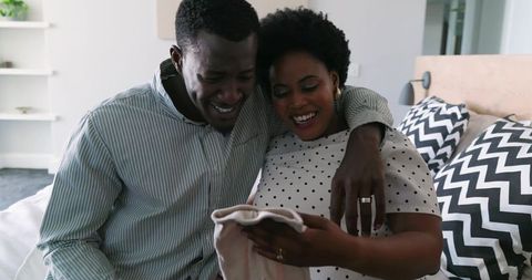 African American Couple Holding Baby Garment on Bed, Smiling and Showing Wedding Rings